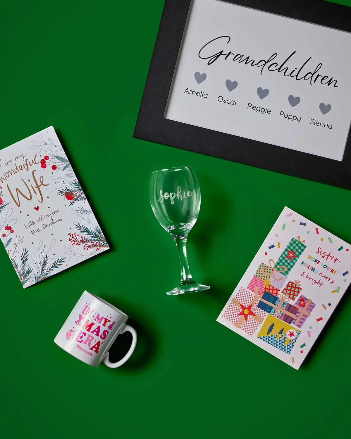Christmas gift collection featuring personalised greeting cards, a framed family print, engraved wine glass, novelty mug, and festive keepsakes on a green background.